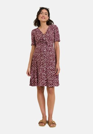 Femme portant une robe bordeaux mi-longue à imprimé floral blanc et manches courtes, associée à des sandales marron et des lunettes de soleil sur la tête.