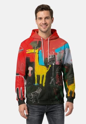 Smiling man wearing colorful hoodie with abstract graffiti art, including a large yellow giraffe and various sketches on red, black, and blue background.