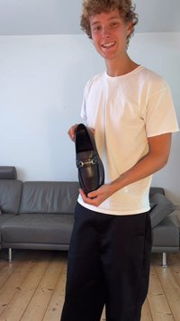 Mocasín de cuero negro con puntera redondeada, con hardware decorativo en plata en la parte frontal, sostenido por una persona con un atuendo casual.
