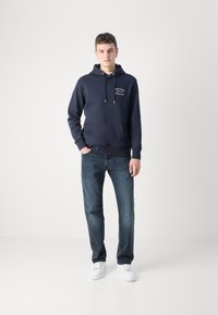Marineblå hettegenser laget av bomullsblanding, med en frontlomme, sølvfargede snorer og hvit tekst. Kombinert med mørkeblå jeans og hvite sneakers.