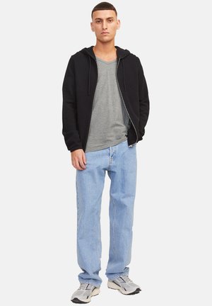 Jonge man staand, gekleed in een zwarte hoodie met rits over een grijs V-hals T-shirt, lichtblauwe spijkerbroek en grijze sneakers, tegen een witte achtergrond.