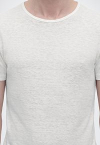 Κοντινό πλάνο ενός ατόμου που φοράει ένα μονόχρωμο, ανοιχτό γκρι, κοντομάνικο μπλουζάκι με στρογγυλή λαιμόκοψη από τους ώμους μέχρι τη μέση.