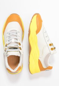 Sneakers with a predominantly white leather upper, orange accents, yellow rubber sole, mesh details, and grey laces. Features metallic eyelets.