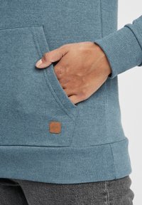 Sudadera azul con bolsillo canguro y puños acanalados. Muestra una etiqueta cuadrada de cuero marrón con texto en la parte inferior izquierda.