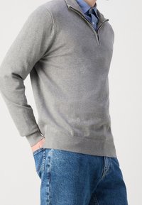 Maglione grigio con zip, maniche lunghe e orlo a coste, indossato sopra una camicia azzurra con colletto e abbinato a jeans di denim blu.