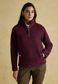 Sieviete, tērpusies rozā krāsas vilnas džemperī ar rāvējslēdzēju un kabatas pogām, kā arī brūnas krāsas ķekars biksēs, stāv uz viegli pelēka fona.