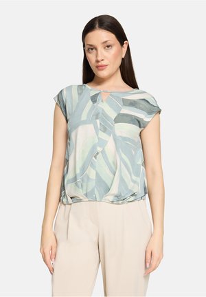 Lightweight blouse featuring a soft blue and green wave pattern, short sleeves, and a keyhole cutout at the neckline. Paired with beige pants.