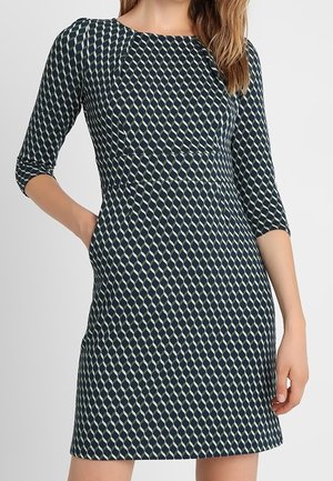 Kobieta ma na sobie dopasowaną sukienkę sięgającą do kolan z rękawami do łokcia, z granatowo-białym geometrycznym wzorem diamentów.