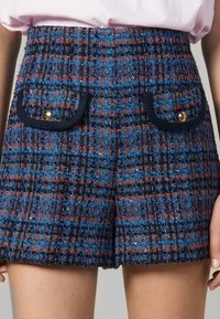 Shorts en tweed taille haute avec un motif à carreaux bleu, noir et rouge, dotés de deux poches à rabat à l'avant avec des bordures noires et des boutons dorés.