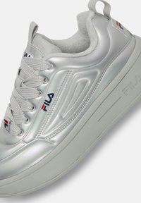 Sneaker sintetiche argento con parte superiore testurizzata, chiusura con lacci, accenti logo e suola in gomma spessa. Include un tassello posteriore per una facile vestibilità.