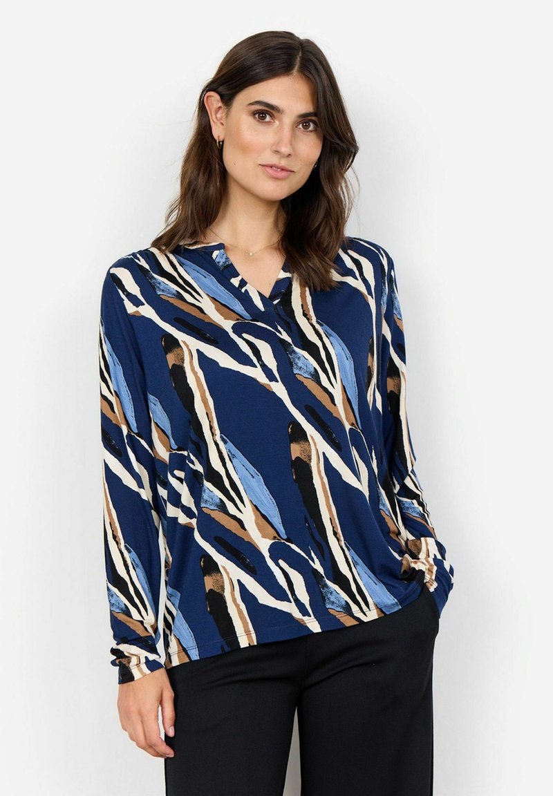 Blusa a maniche lunghe blu navy con motivi astratti blu, beige e neri. Design a V, tessuto morbido e vestibilità rilassata. Abbinata a pantaloni neri.