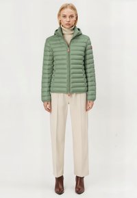 Lichtgroene pufferjack met horizontale gestikte textuur, kap en oranje ritslus. Draag het over een beige turtleneck en crèmekleurige broek.