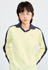 Polo-shirt à manches longues Puma en jaune clair avec des accents bleu marine, présentant une texture côtelée et un design en V. Logo discret sur la poitrine.