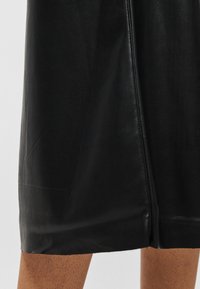 Melnas ādas svārki ar centra aizmugures šuvi un nelielu šķēlumu virs ceļgaliem smilškrāsas ādas fona priekšā.