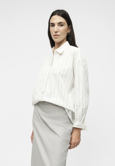 Γυναίκα με μακριά σκουρόχρωμα μαλλιά, που φοράει ασημένια κρίκους μανικετόκουμπα, ένα άσπρο πουκάμισο με λεπτές κάθετες ρίγες και ένα ανοιχτό γκρι φόρεμα.
