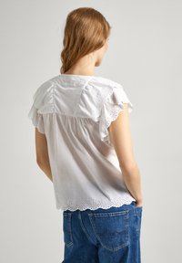 Hvid bomuldsbluse med volangærmer og bølget hæklet kant. Båret med højtaljede blå jeans, der fremhæver et afslappet fit design.