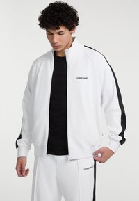 Witte zip-up jas met zwarte zijstrepen en logodetail. Zachte stof met een gladde textuur. Gecombineerd met een zwart T-shirt met ronde hals.