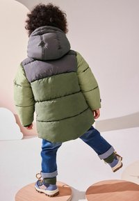 Chaqueta acolchada verde y gris con capucha, con secciones acolchadas horizontales, combinada con unos vaqueros azules y botas resistentes.