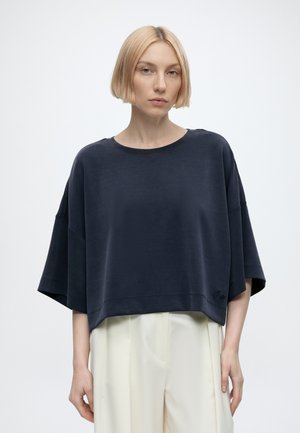 Blond kvinde med kort bob-frisure iført en løs, mørk marineblå oversize kortærmet top og højtaljede cremefarvede bukser, stående mod en hvid baggrund.