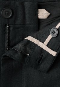Gros plan sur la ceinture d'un pantalon en tissu noir avec des boutons noirs, une fermeture éclair et des détails de couture beige à l'intérieur de la ceinture.