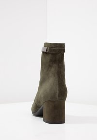 Botín de ante verde oliva hasta el tobillo con punta redonda, tacón ancho y detalle de correa ajustable en el cuello. Textura suave con acentos mínimos.