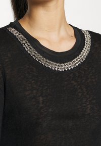 Schwarzer Pullover mit transparenter Textur, der einen mit Metallstiften besetzten Halsausschnitt mit einem dekorativen Muster akzentuiert. Klassisches Rundhalsdesign.