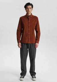Braune Corduroy-Button-Up-Hemd mit Kragen, kombiniert mit grauen schmalen Hosen und schwarzen Sneakers, stehend vor einem neutralen Hintergrund.
