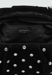 Interior de bolso de terciopelo negro con tachuelas plateadas distribuidas uniformemente. Presenta un forro de tela negra y una etiqueta metálica de la marca etiquetada como "MNG".