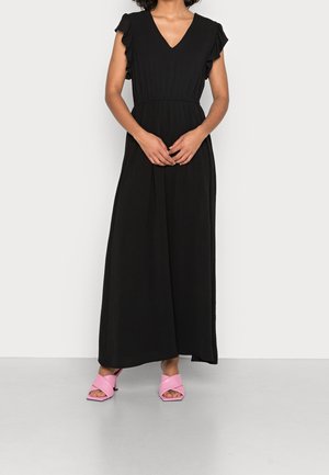 Kvinde iført en lang sort kjole med korte flæseærmer og pink hælede sandaler, stående mod en ensfarvet hvid baggrund.