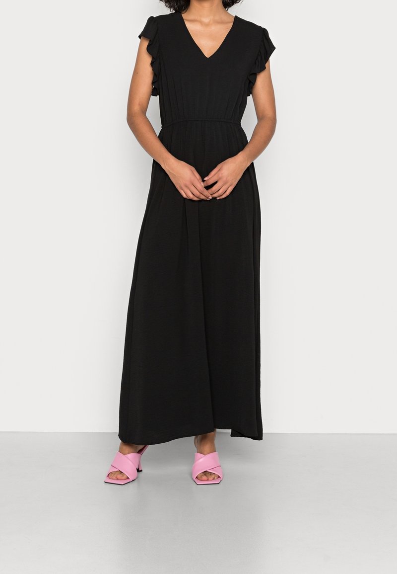 Femme portant une longue robe noire avec des manches courtes volantées et des sandales roses à talons, debout sur un fond blanc uni.