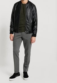 Homme portant une veste en cuir noire, un pull en tricot vert olive, un pantalon gris avec une bande latérale, et des baskets noires avec des semelles blanches.