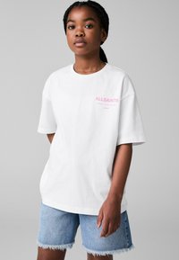 Balta per didelė t-shirt su rožiniu "ALLSAINTS" logotipu, derinama su šviesiai mėlynomis džinsinėmis šortais su išdraskytu kraštu. Lygi medžiaga, laisvas siluetas.