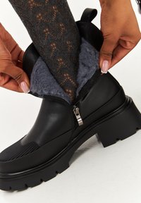 Bottine noire avec une finition texturée, fermeture éclair, doublure en fourrure grise et semelle en caoutchouc épaisse. Une main soulève la doublure en fourrure.