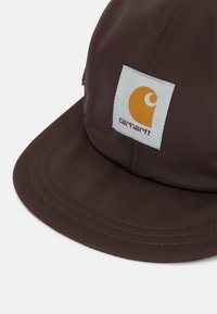 Cappello marrone realizzato in un tessuto resistente, con visiera piatta e una toppa rettangolare con un design giallo. La cucitura è visibile lungo le cuciture.