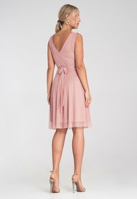 Różowa, bezrękawna sukienka z dekoltem w kształcie litery V na plecach i wiązaniem w pasie. Spódnica ma warstwowy, zwiewny krój. Noszona z sandałami na obcasie.