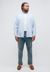 Άνδρας με πλεγμένα μαλλιά, φοράει ανοιχτό μπλε ριγέ πουκάμισο πάνω από λευκή μπλούζα, μπλε τζιν και καφέ μοκασίνια, στέκεται και κοιτάζει δεξιά.