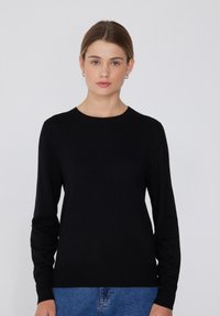Schwarzer langärmliger Pullover aus weichem Strickmaterial mit Rundhalsausschnitt, schlanker Passform und gerippten Bündchen und Saum, getragen über blauen Jeans.