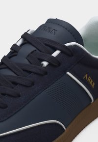 Κοντινό πλάνο αθλητικού παπουτσιού σε ναυτικό μπλε με μαύρα κορδόνια, λευκή επένδυση, πλευρικό πάνελ με τρύπες και χρυσό λογότυπο "ARKK" στο πλάι και στη γλώσσα.