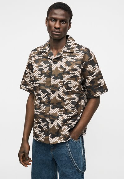 Νεαρός άνδρας με κοντομάνικο πουκάμισο με καμουφλάζ και μπλε τζιν με αλυσίδα, που στέκεται μπροστά σε ένα απλό φόντο.