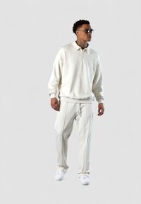 Weißer Fleece-Zip-Up-Pullover mit Kragen, kombiniert mit passenden, schmal geschnittenen Jogginghosen. Trägt weiße Sneakers und schwarze Sonnenbrille.