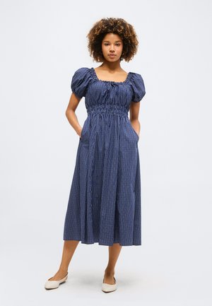 Γυναίκα που στέκεται με τα χέρια στις τσέπες φορώντας ένα μπλε καρό μίντι φόρεμα με φουσκωτά μανίκια και λευκά επίπεδα παπούτσια.