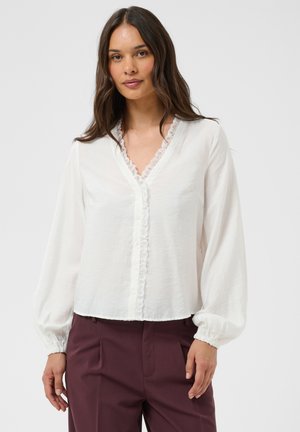 Femme aux longs cheveux bruns porte un chemisier blanc à manches longues avec bordure en dentelle et un pantalon taille haute bordeaux, debout devant un fond neutre.