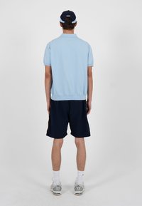Homme portant un polo à manches courtes bleu clair, un short bleu marine avec des bandes latérales, des chaussettes blanches, des baskets, et une casquette de baseball bleu marine, debout de dos.