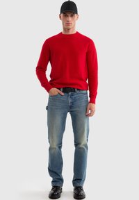 Czerwony sweter z dzianiny, niebieskie dżinsy, czapka w kolorze czarnym, czarny pasek, czarne buty. Sweter ma okrągły dekolt; dżinsy mają lekkie przetarcia na kolanach.