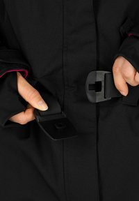 Black fabric outerwear featuring adjustable straps and buckles. Close-up on hands fastening the straps. Interior has pink accents.