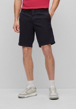 Hombre con pantalones cortos negros hasta la rodilla, calcetines blancos, zapatillas grises y blancas y una camiseta roja con una mano en el bolsillo.