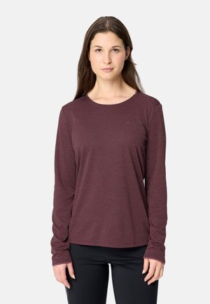 Frau mit glattem braunem Haar, die ein langärmeliges weinrotes Shirt und schwarze Hose trägt, steht vor einem schlichten hellen Hintergrund.