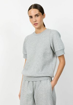 Femeie purtând un hanorac gri deschis cu mâneci scurte și mansete și pantaloni de trening gri asortați, stând pe un fundal alb simplu.