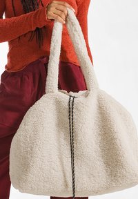 Crèmekleurige textiel tas met een fluffy textuur, voorzien van twee stevige handvatten en een contrasterende zwarte koorddetail langs het midden.