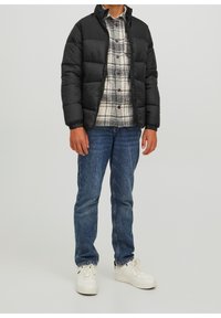 Osoba w czarnej kurtce puchowej, w kratkowanej koszuli, niebieskich dżinsach i białych trampkach stoi na jednolitym jasnym tle.